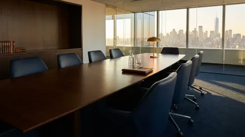 Empty executive boardroom at golden hour — polished mahogany table, deep-navy leather chairs, glass wall with city dusk skyline